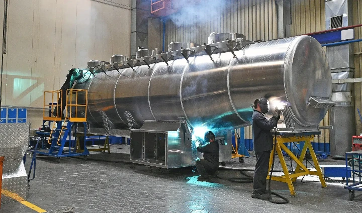 Tres soldadores con equipo de protección trabajan en un gran tanque de acero inoxidable sobre soportes amarillos en un gran taller industrial con humo y destellos de soldadura.